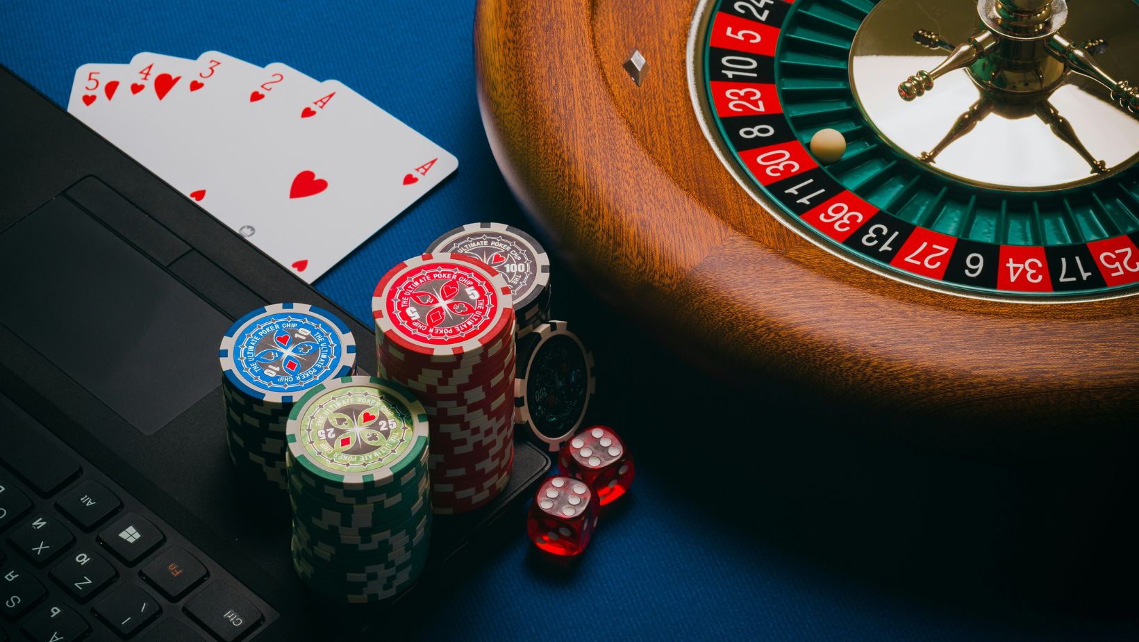 A roulette wheel, playing cards, poker chips, and dice next to a laptop